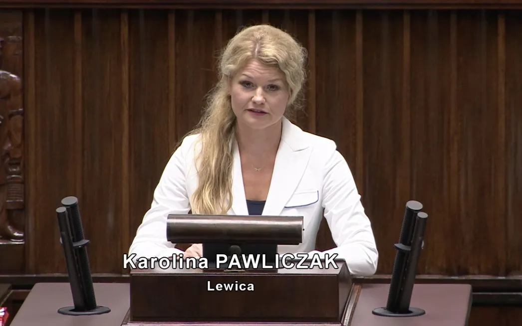 Kobieta w białej marynarce przemawia z mównicy podczas pilnej interpelacji w parlamencie.