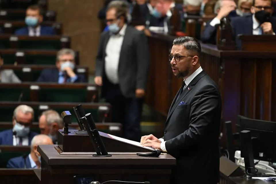 Mężczyzna w garniturze przemawia z mównicy podczas posiedzenia w sali plenarniej, trzymając dokumenty, podczas gdy w tle siedzący uczestnicy w maseczkach obserwują wydarzenie.