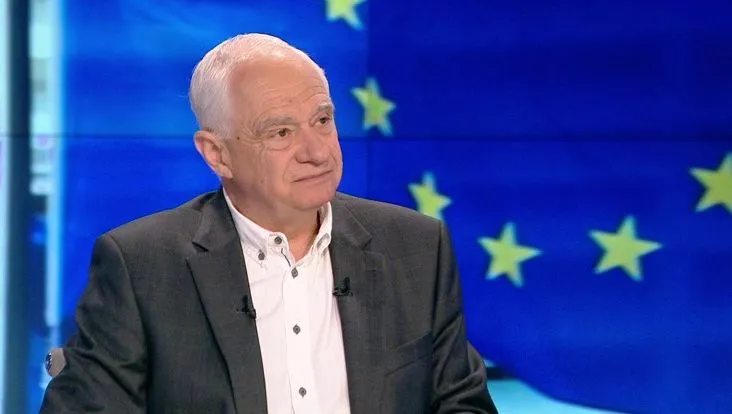 Mężczyzna w garniturze siedzi w studiu, a w tle widać niebieską flagę Unii Europejskiej z żółtymi gwiazdami.