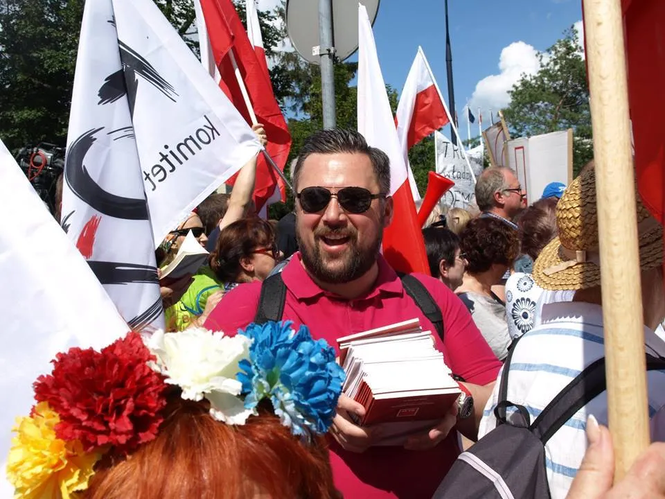 Uśmiechnięty mężczyzna w różowej koszulce i okularach przeciwsłonecznych rozdaje pakiety materiałów w tłumie demonstrantów z flagami.