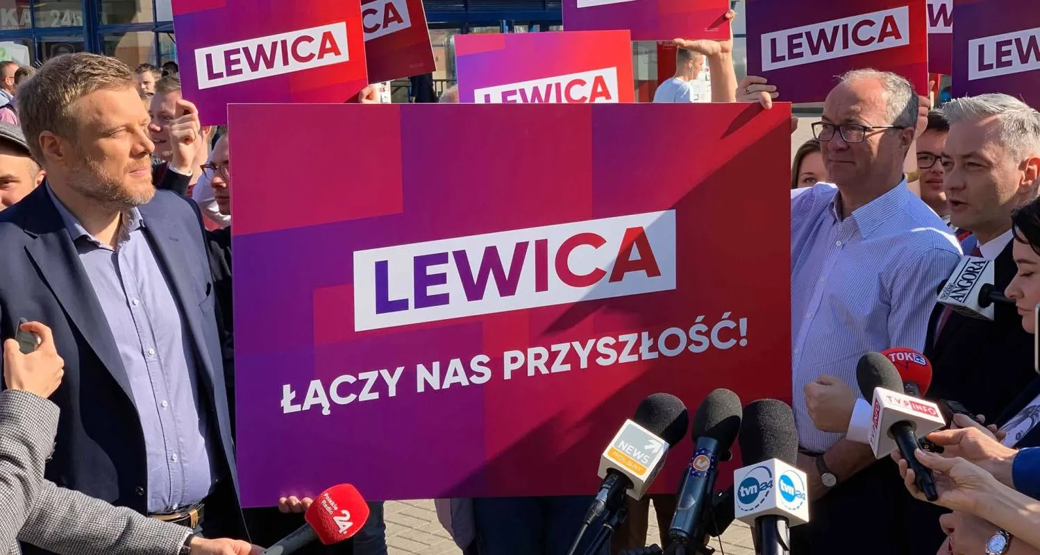 Grupa zwolenników Lewicy trzyma duży, kolorowy plakat z napisem LEWICA i hasłem „Łączy nas przyszłość!”, podczas konferencji prasowej z mikrofonami.
