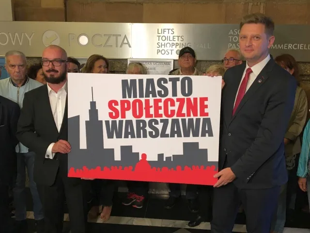 Grupa ludzi trzyma plakat z napisem Miasto Społeczne Warszawa, na którym widoczna jest czarna sylweta miasta na czerwonym tle.