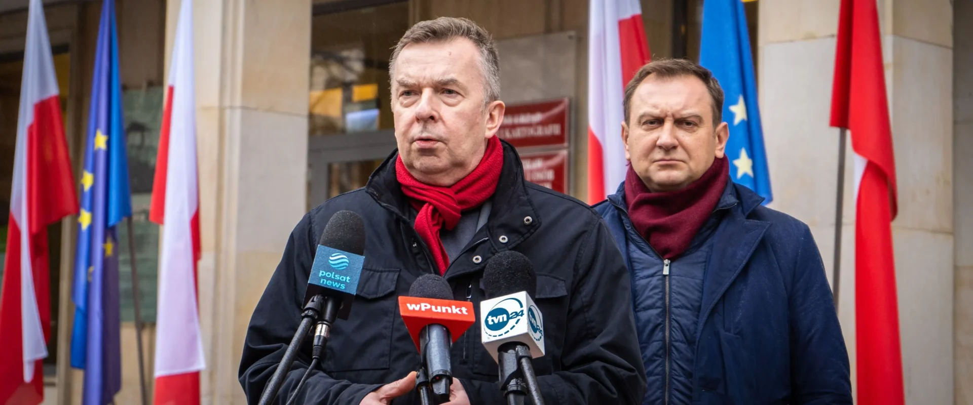 Na zdjęciu widać dwóch mężczyzn przemawiających podczas konferencji prasowej, stojących przed grupą flag, w tym flagami Polski i Unii Europejskiej.