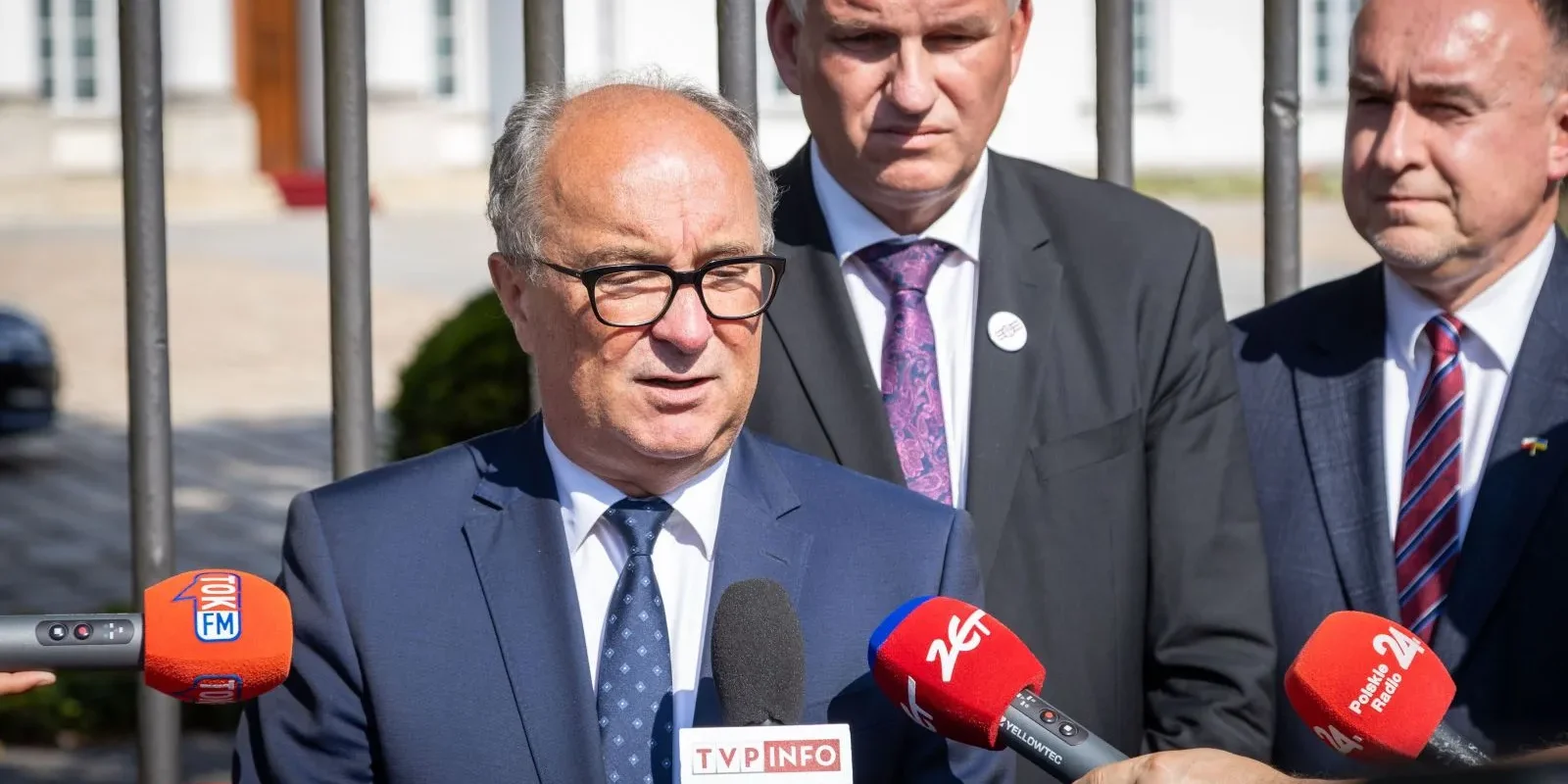 Mężczyzna w granatowym garniturze i okularach udziela wywiadu przed mikrofonami dziennikarzy podczas konferencji prasowej na zewnątrz.