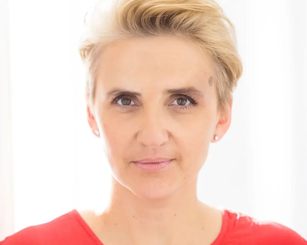 Portret kobiety w czerwonej koszuli, z krótkimi, jasnymi włosami, patrzącej prosto w obiektyw na tle jasnego okna.