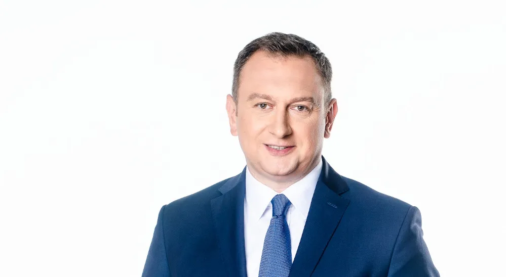 Portret mężczyzny w granatowym garniturze, białej koszuli i niebieskim krawacie, na białym tle, uśmiechnięty do kamery.