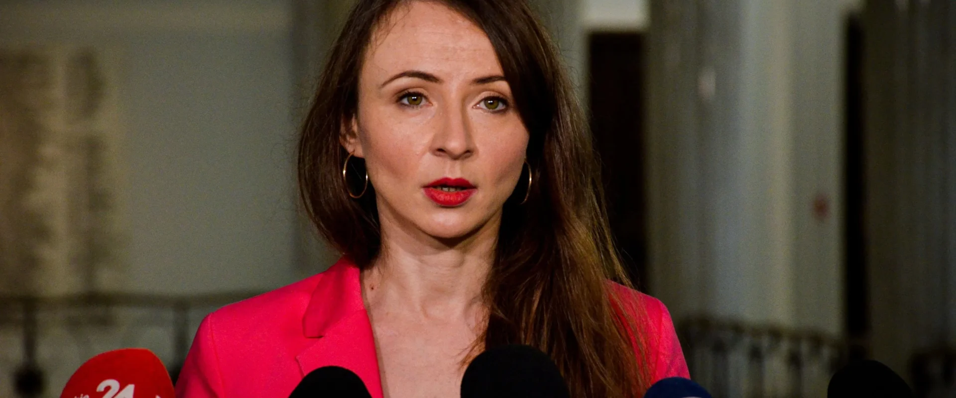 Kobieta w różowej marynarce przemawia do mediów podczas konferencji prasowej, stojąc przed kilkoma mikrofonami.