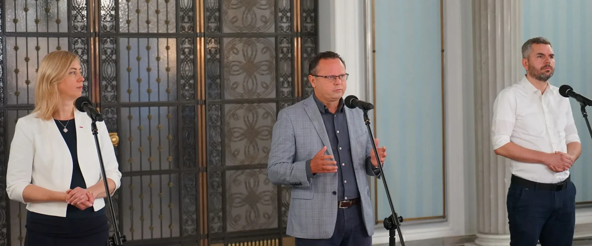 Trzy osoby stojące przy mikrofonach podczas konferencji prasowej w eleganckiej sali z ozdobnymi drzwiami w tle.