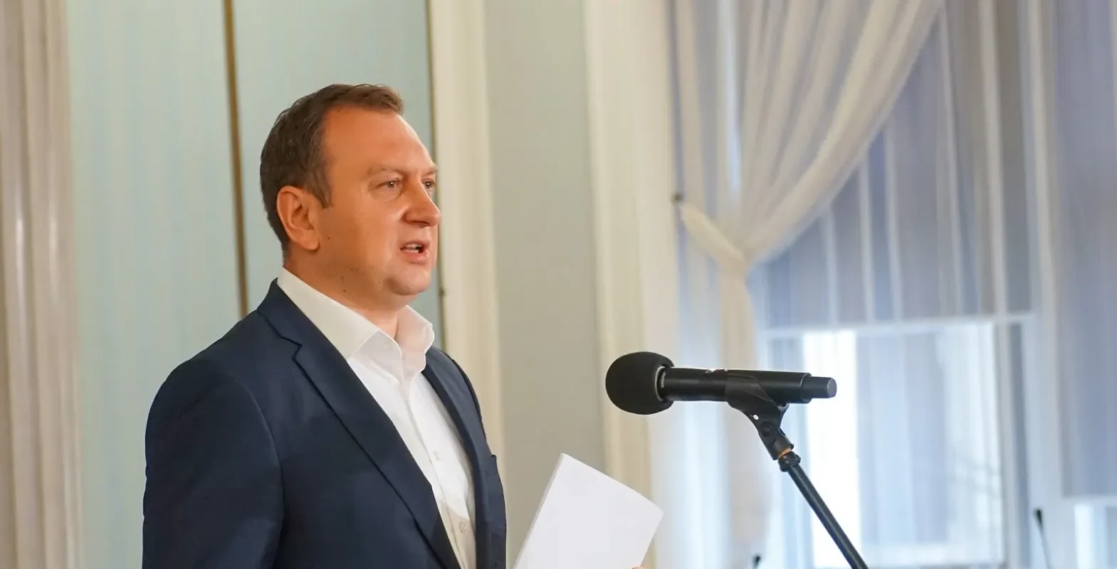 Mężczyzna w garniturze przemawia do mikrofonu, trzyma kartkę w dłoni podczas konferencji.