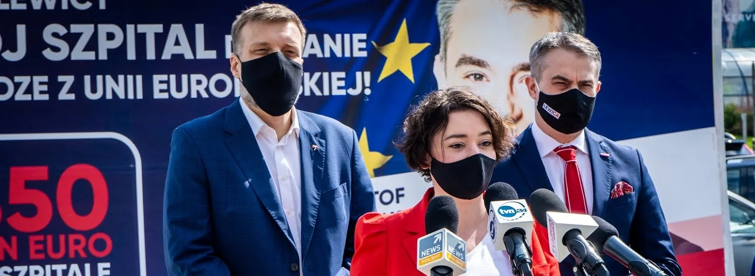 Trzy osoby w maseczkach stoją przed banerem Lewicy z mikrofonami podczas konferencji prasowej, a kobieta w czerwonej marynarce jest na pierwszym planie.