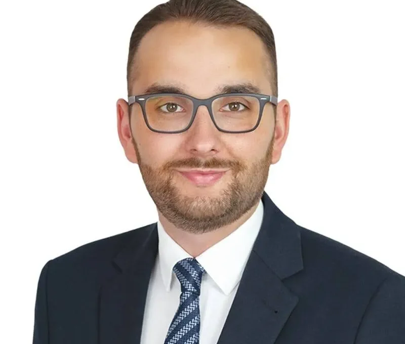Portret mężczyzny w garniturze, białej koszuli i krawacie, noszącego okulary, na neutralnym białym tle.