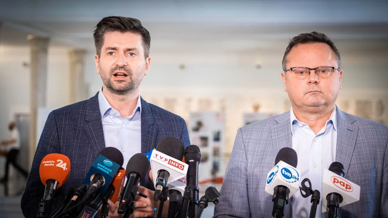 Dwóch mężczyzn w garniturach przemawia podczas konferencji prasowej, stojąc przed zestawem mikrofonów w sprawie uchodźców w Polsce.