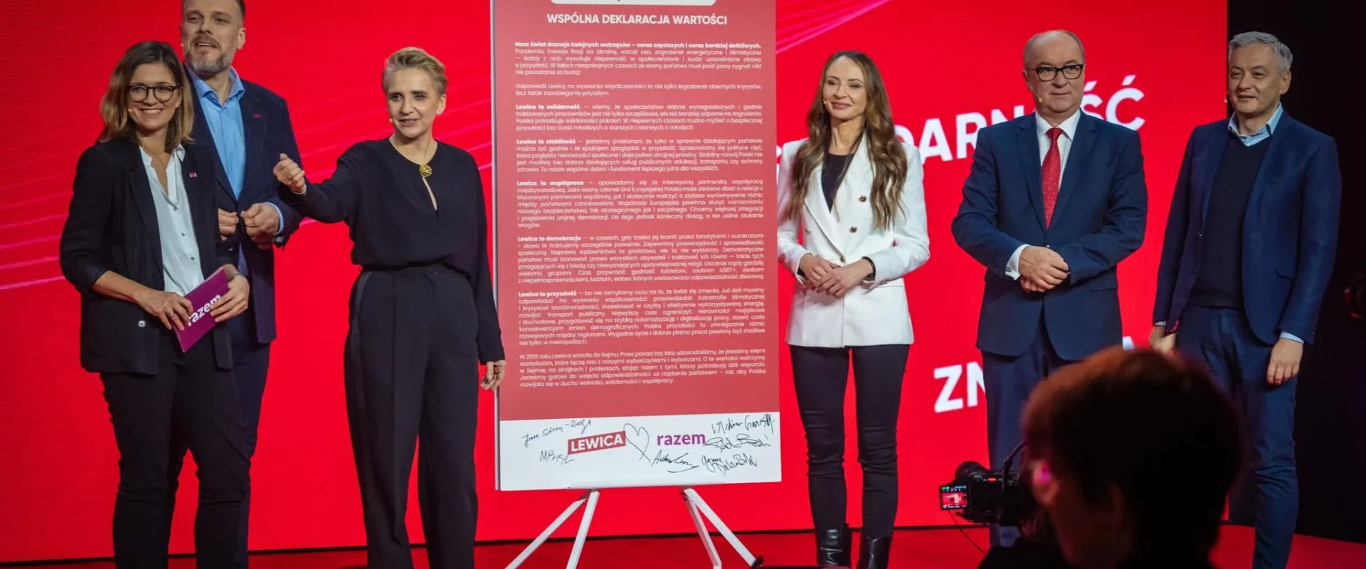 Na zdjęciu widoczna jest grupa polityków podczas wydarzenia promującego deklarację wartości, z czerwonym tłem i dużym plakatem z napisem "Czas wyprowadzić Polskę z kryzysu".