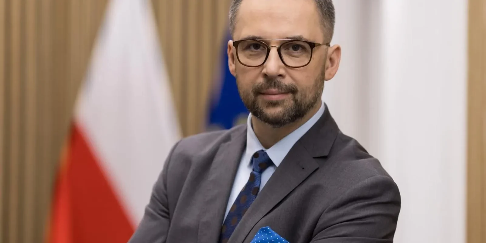 Mężczyzna w szarym garniturze, okularach i niebieskim krawacie z poszetką stoi z założonymi rękami na tle flag.