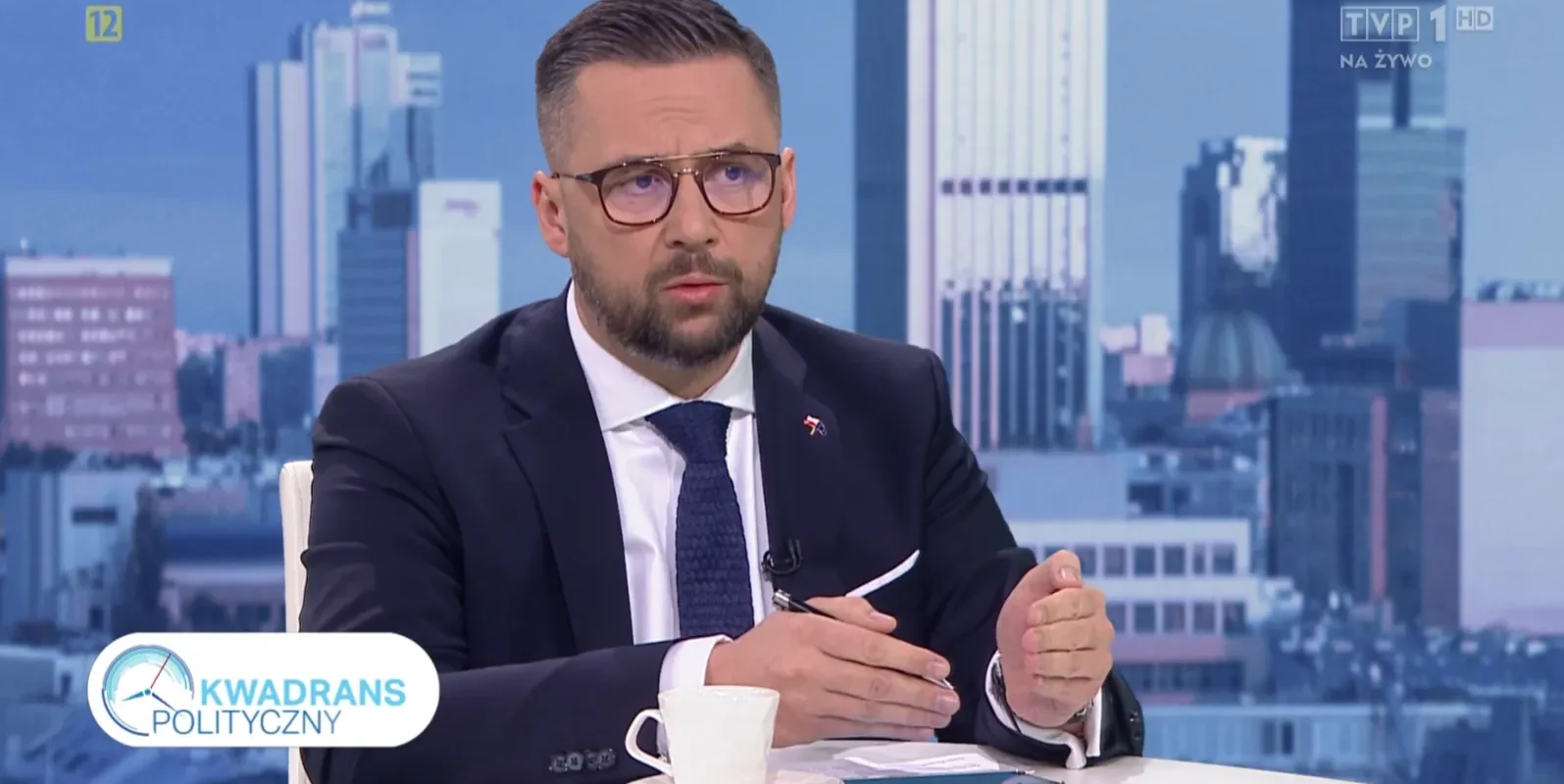 Mężczyzna w eleganckim garniturze siedzi przy stole w studiu telewizyjnym, rozmawiając na temat polityki, z nowoczesnym miastem w tle.