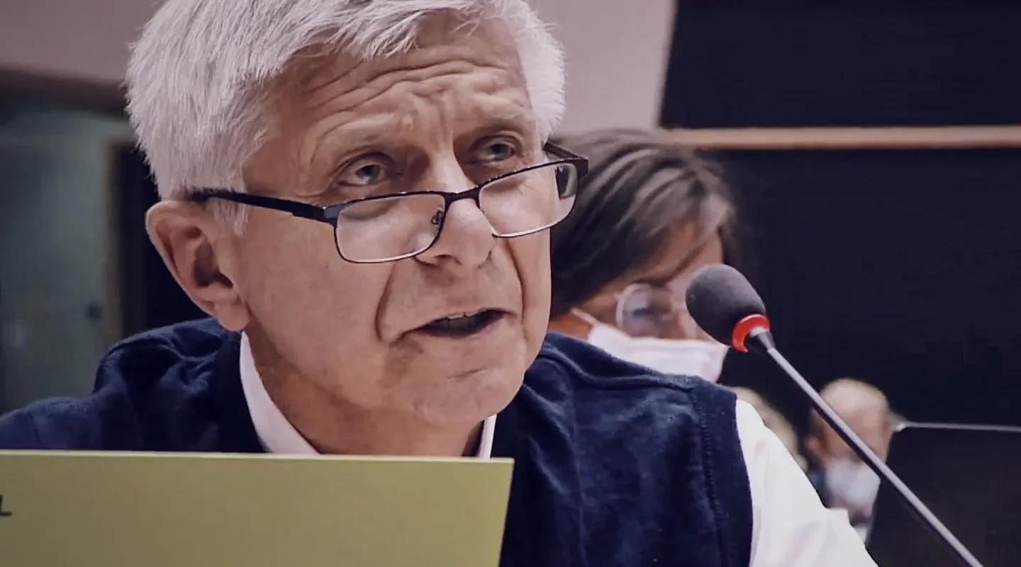 Starszy mężczyzna z siwymi włosami i okularami siedzi przy mikrofonie podczas konferencji, trzyma żółtą kartkę.