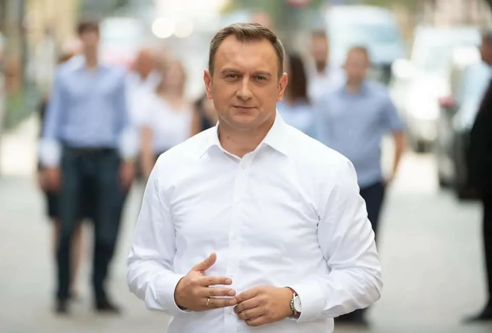 Mężczyzna w białej koszuli stoi na ulicy, a w tle widoczni są rozmyci przechodnie.