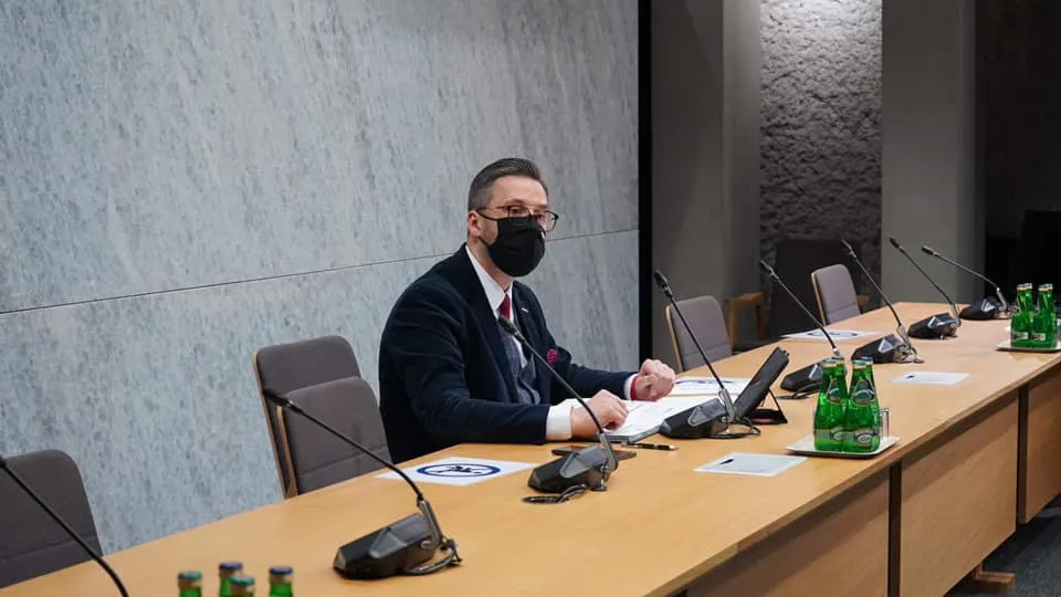 Osoba w garniturze i maseczce siedzi przy długim stole konferencyjnym z mikrofonami i zielonymi butelkami wody.