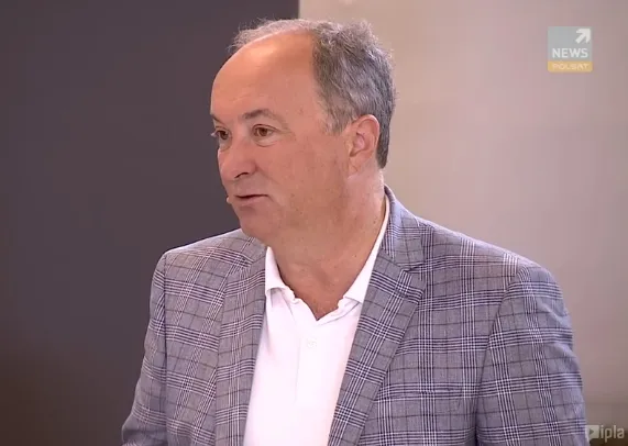 Mężczyzna w szarej kratkowanej marynarce i białej koszuli rozmawia w studiu telewizyjnym.