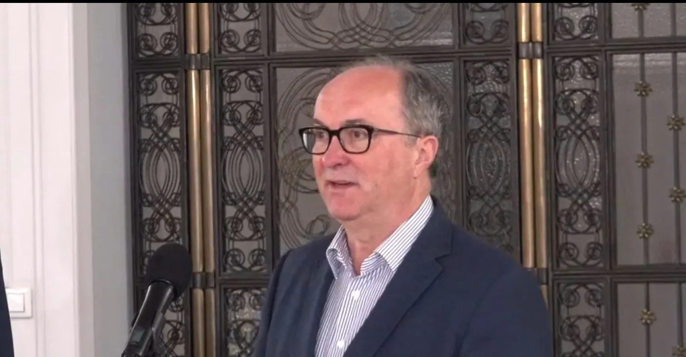 Mężczyzna w garniturze i okularach przemawia do mikrofonu podczas konferencji prasowej, stojąc przed ozdobnymi, kutymi drzwiami.