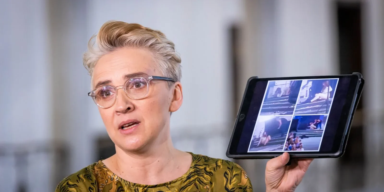 Kobieta w okularach trzyma tablet, na którym wyświetlone są zdjęcia, podczas gdy wyraża emocje podczas rozmowy.
