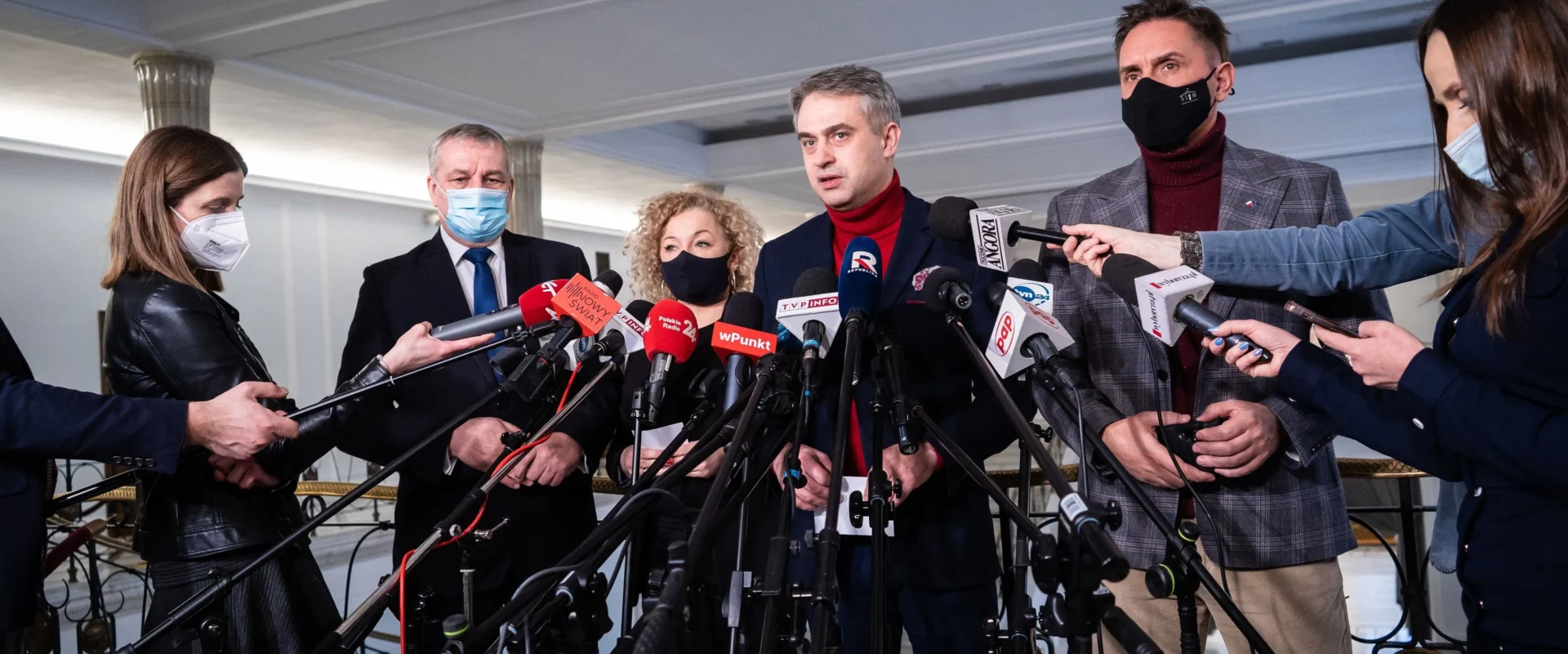 Grupa polityków i dziennikarzy podczas konferencji prasowej, otoczona licznymi mikrofonami i kamerami, wszyscy w maseczkach.