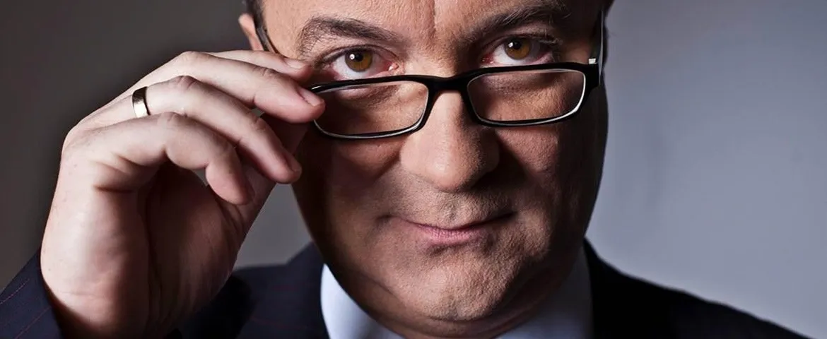 Zbliżenie na mężczyznę w okularach i garniturze, lekko unoszącego oprawki i patrzącego prosto w obiektyw.
