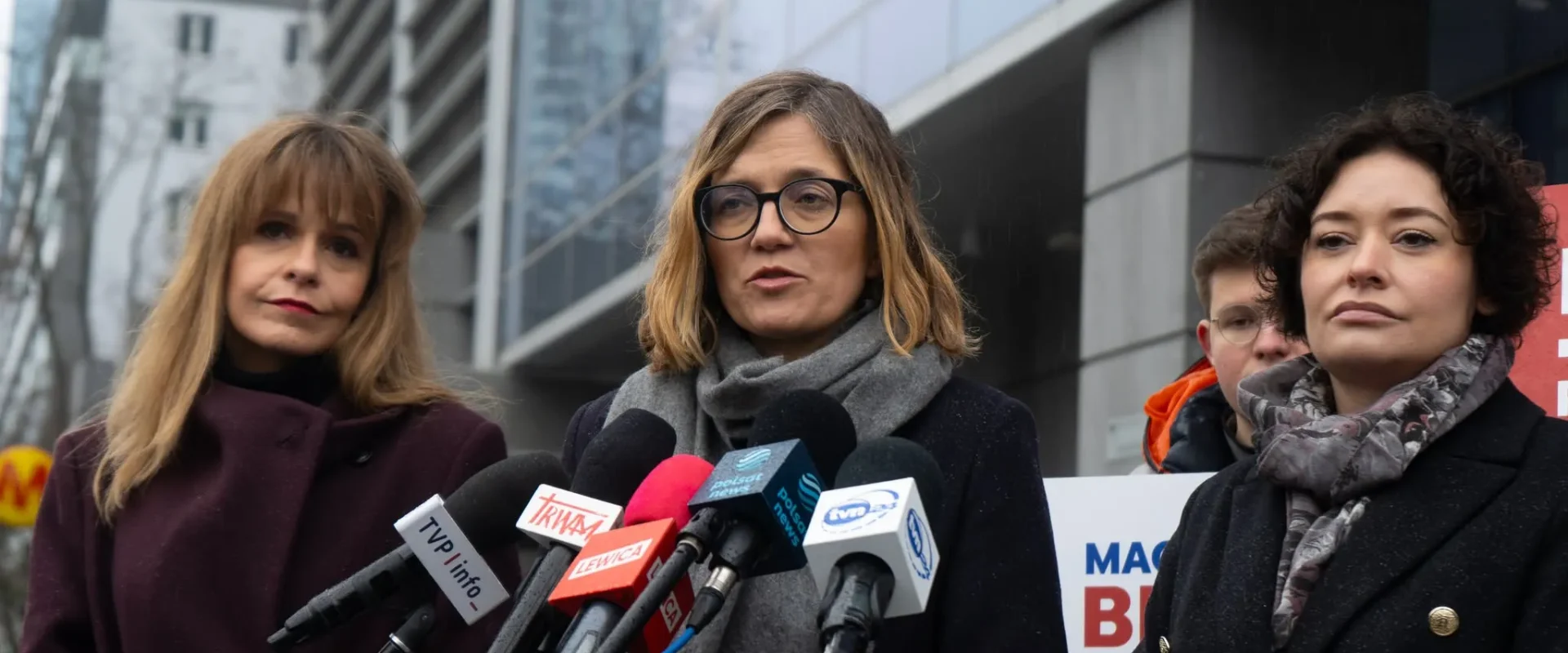 Trzy kobiety stoją przy zestawie mikrofonów podczas konferencji prasowej na zewnątrz nowoczesnego budynku, z których jedna wygłasza oświadczenie.