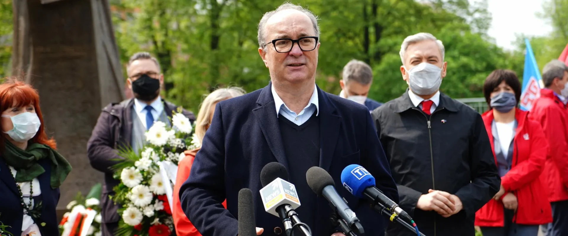 Centralnie ustawiony mężczyzna w granatowym garniturze przemawia do mikrofonów podczas obchodów 1 maja, otoczony przez innych uczestników z kwiatami i flagami w parku.