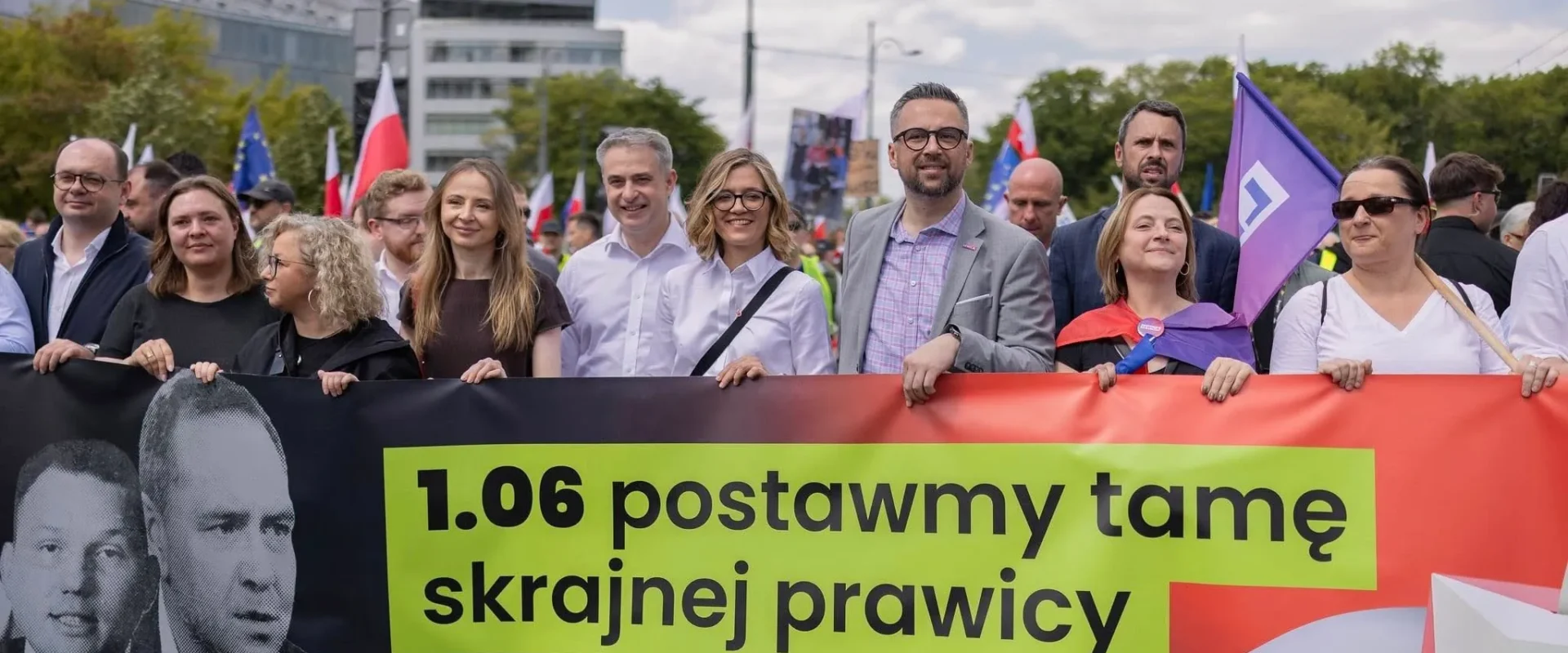 Grupa uczestników Wielkiego Marszu Patriotów trzyma na tle budynków duży baner z napisem „1.06 postawmy tamę skrajnej prawicy” oraz widocznymi flagami.