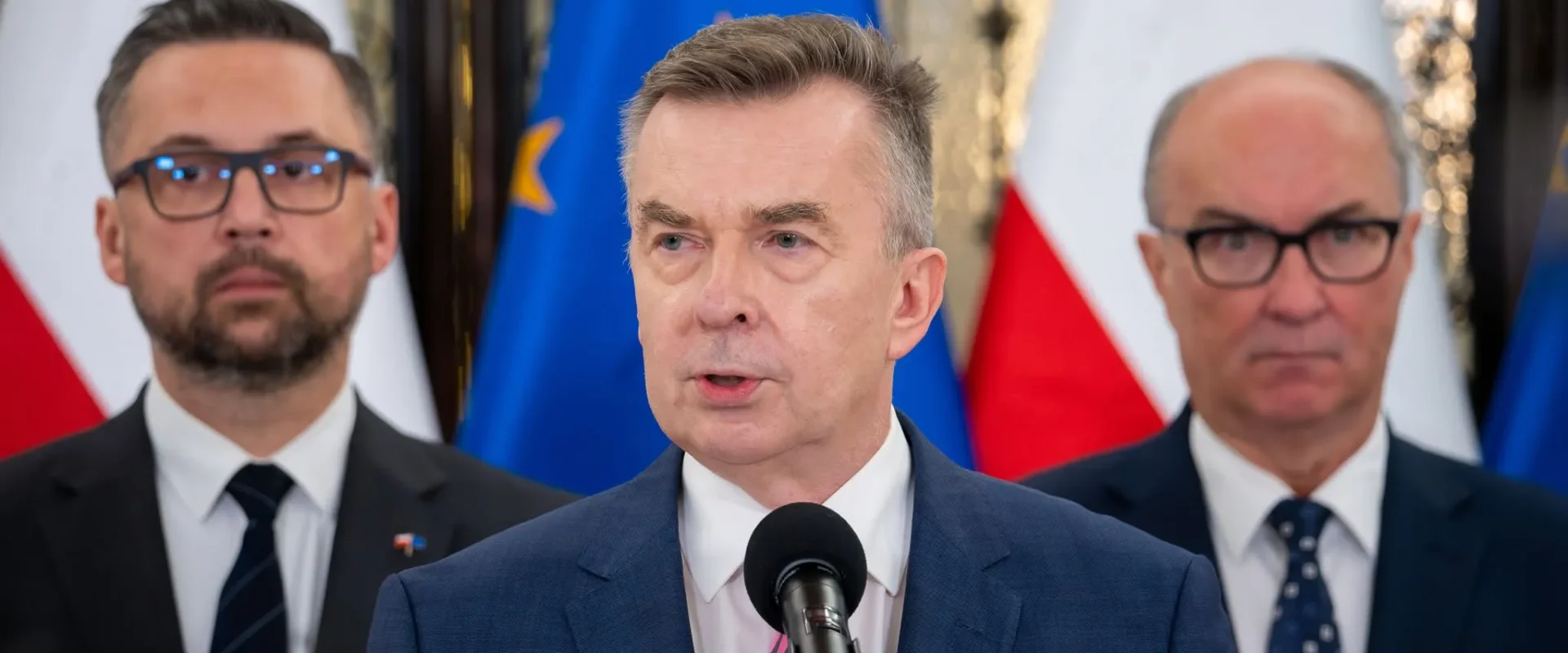 Mężczyzna w granatowym garniturze przemawia do mikrofonu podczas konferencji prasowej, za nim stoją dwie osoby, a w tle widać flagi Polski i Unii Europejskiej.