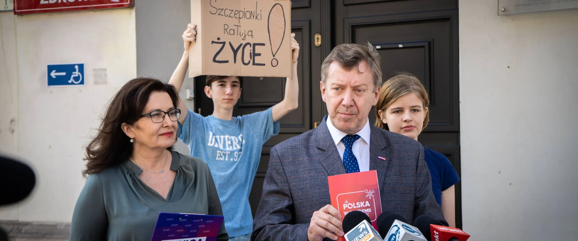 Mężczyzna w garniturze przemawia do mikrofonów przed wejściem do budynku Ministerstwa Zdrowia; za nim osoba trzyma karton z napisem „Szczepionki ratują ŻYCIE!”, a obok stoi kobieta z teczką Lewica.