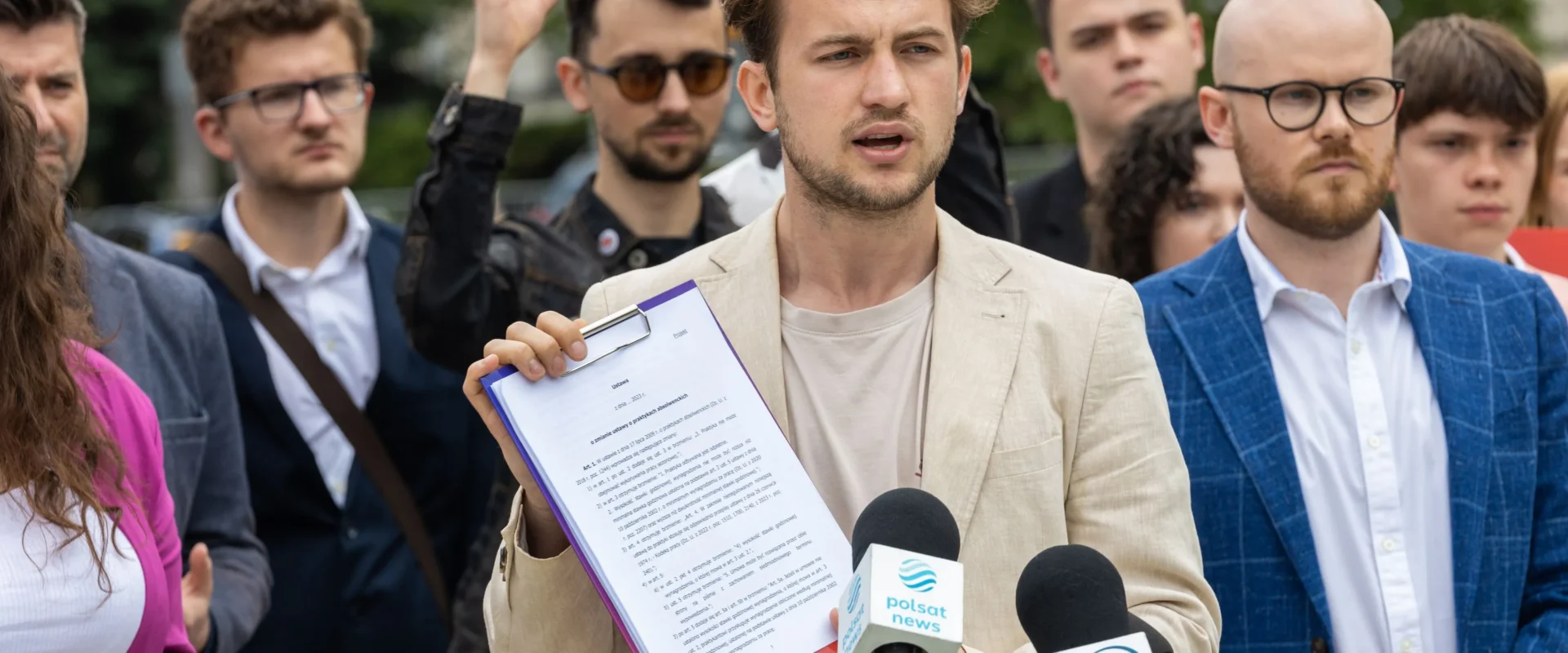 Mężczyzna w jasnobeżowej marynarce trzyma teczkę z dokumentem i przemawia do mikrofonów podczas konferencji prasowej, otoczony grupą osób.