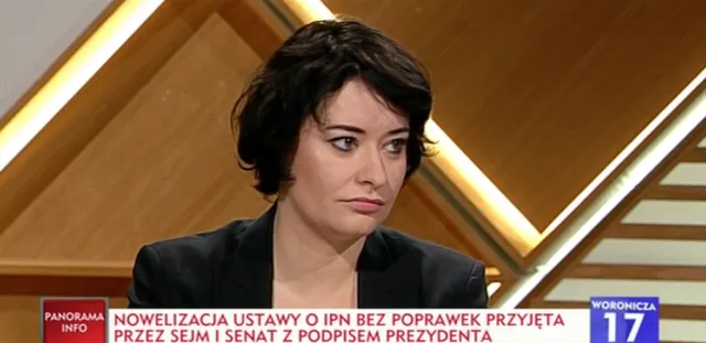 Kobieta w czarnej marynarce w studiu telewizyjnym patrzy na bok ze skupionym, poważnym wyrazem twarzy.