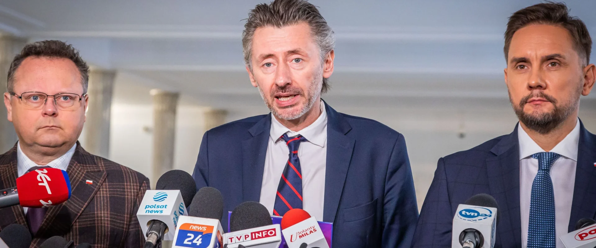 Na zdjęciu trzej mężczyźni stoją przed mikrofonami podczas konferencji prasowej w jasnym wnętrzu.