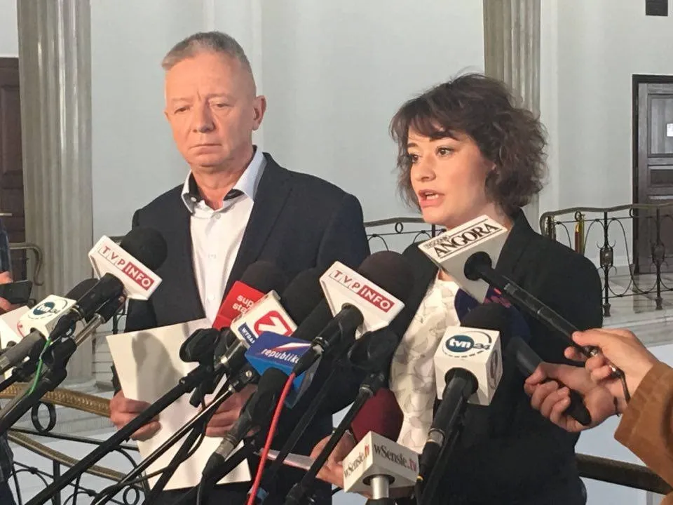 Dwie osoby przemawiają do mediów przy stojaku z licznymi mikrofonami w korytarzu Sejmu.