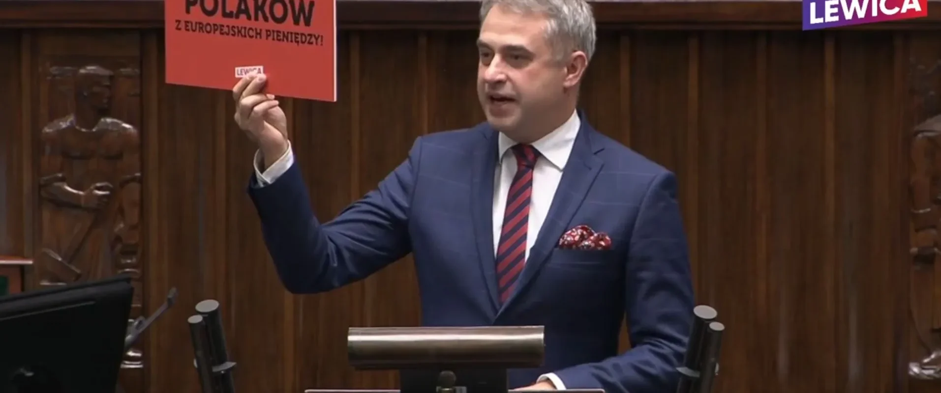 Polityk trzyma w ręku kartkę z napisem "PIS OKRADA POLAKÓW Z EUROPEJSKICH PIENIĘDZY!" podczas wystąpienia w Sejmie.