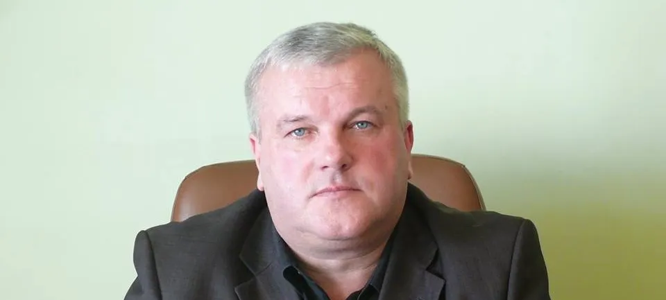 Portret mężczyzny w czarnym garniturze, siedzącego na brązowym fotelu, na jasnym zielonkawym tle.