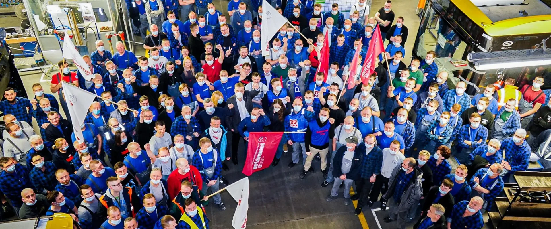 Grupa pracowników w fabryce, ubranych w niebieskie koszulki i maski, z flagami i transparentami, gromadzi się w hali produkcyjnej.