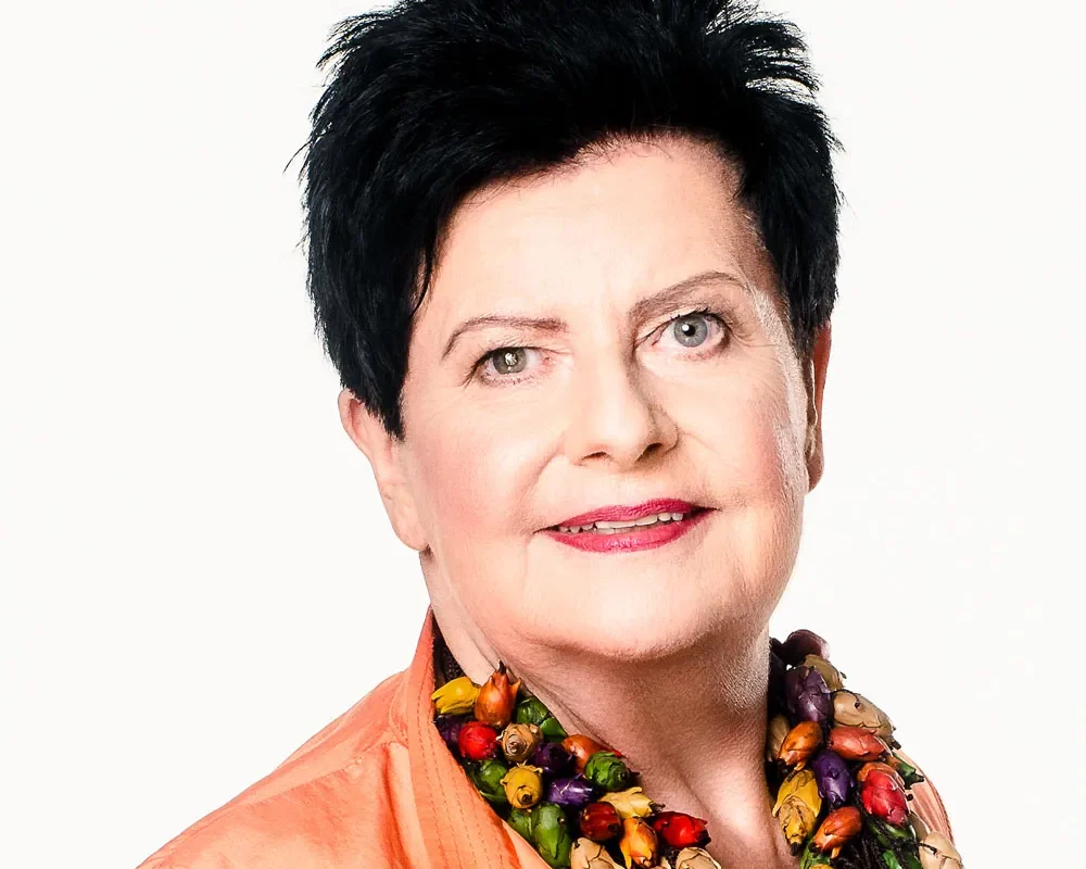 Portret kobiety z krótkimi czarnymi włosami, uśmiechniętej, w pomarańczowej bluzce i kolorowej koralikowej biżuterii na białym tle.