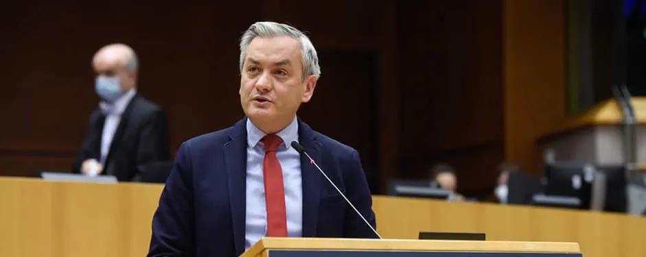 Na zdjęciu przedstawiony jest mężczyzna przemawiający z mównicy podczas sesji w sali obrad, z innymi osobami w tle.