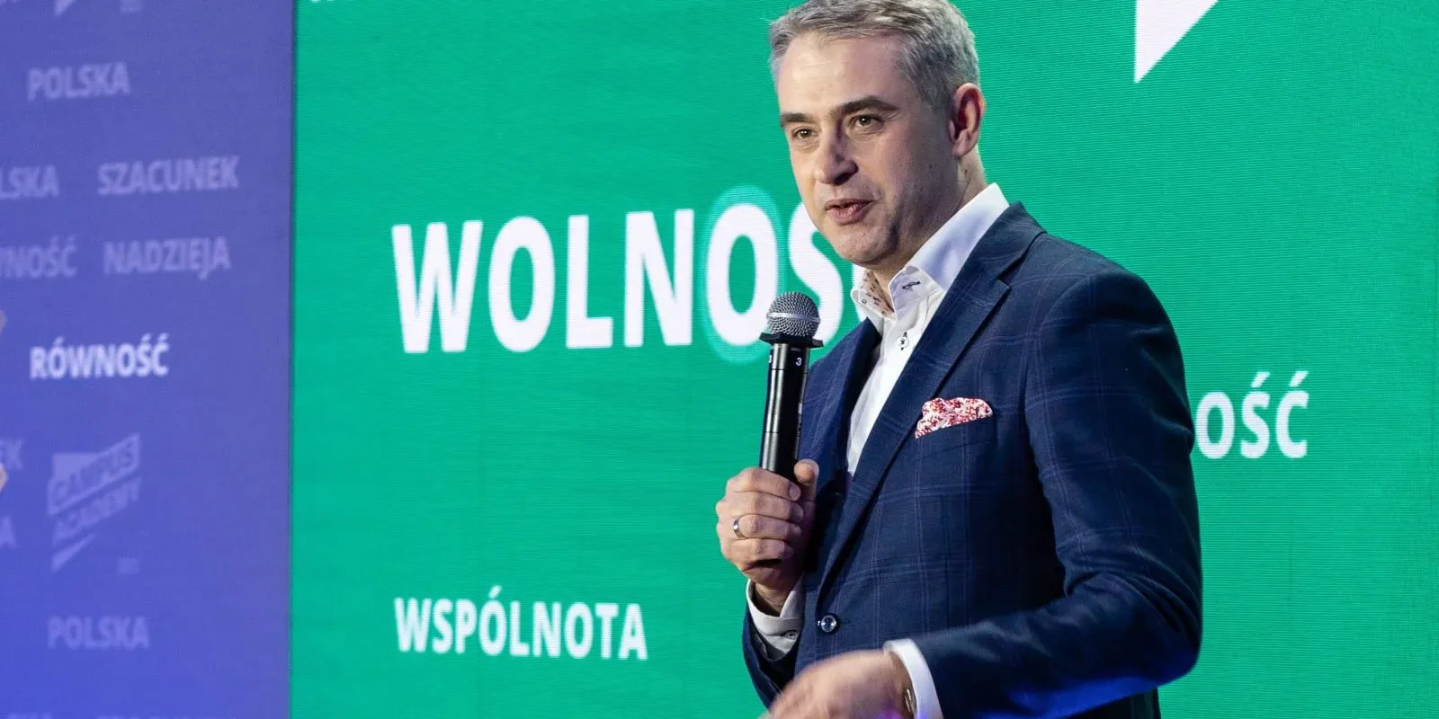 Mężczyzna w granatowym garniturze trzyma mikrofon na scenie, na tle zielonego ekranu z napisami „WOLNOŚĆ” i hasłami „WSPÓLNOTA” oraz „RÓWNOŚĆ”.