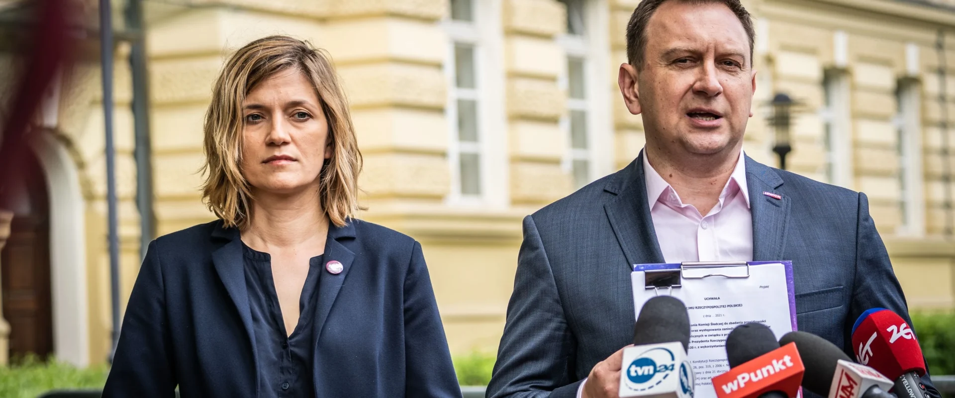 Dwóch polityków lewicy stoi przed budynkiem podczas konferencji prasowej; mężczyzna po prawej trzyma dokument i przemawia do mikrofonów, obok stoi kobieta.