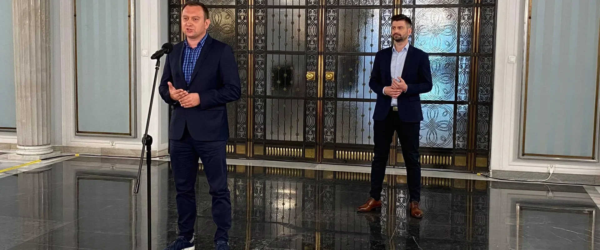 Dwóch mężczyzn w garniturach stoi w eleganckim holu z ozdobnymi drzwiami w tle, jeden przemawia do mikrofonu.