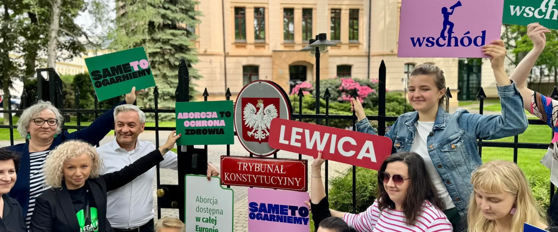 Grupa osób stojąca przy ogrodzeniu przed budynkiem trzyma kolorowe plakaty popierające Lewicę i europejską inicjatywę obywatelską „My voice my choice”.