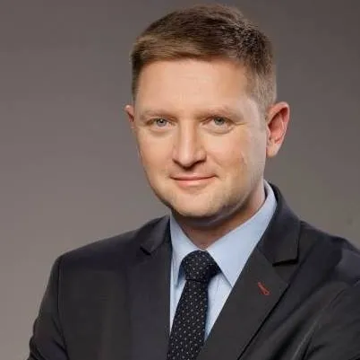 Portret uśmiechniętego mężczyzny w garniturze i krawacie na jednolitym, jasnoszarym tle.