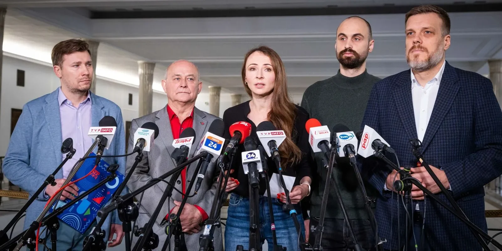 Na zdjęciu widoczna jest grupa pięciu osób stojących przed mikrofonami, podczas konferencji prasowej.