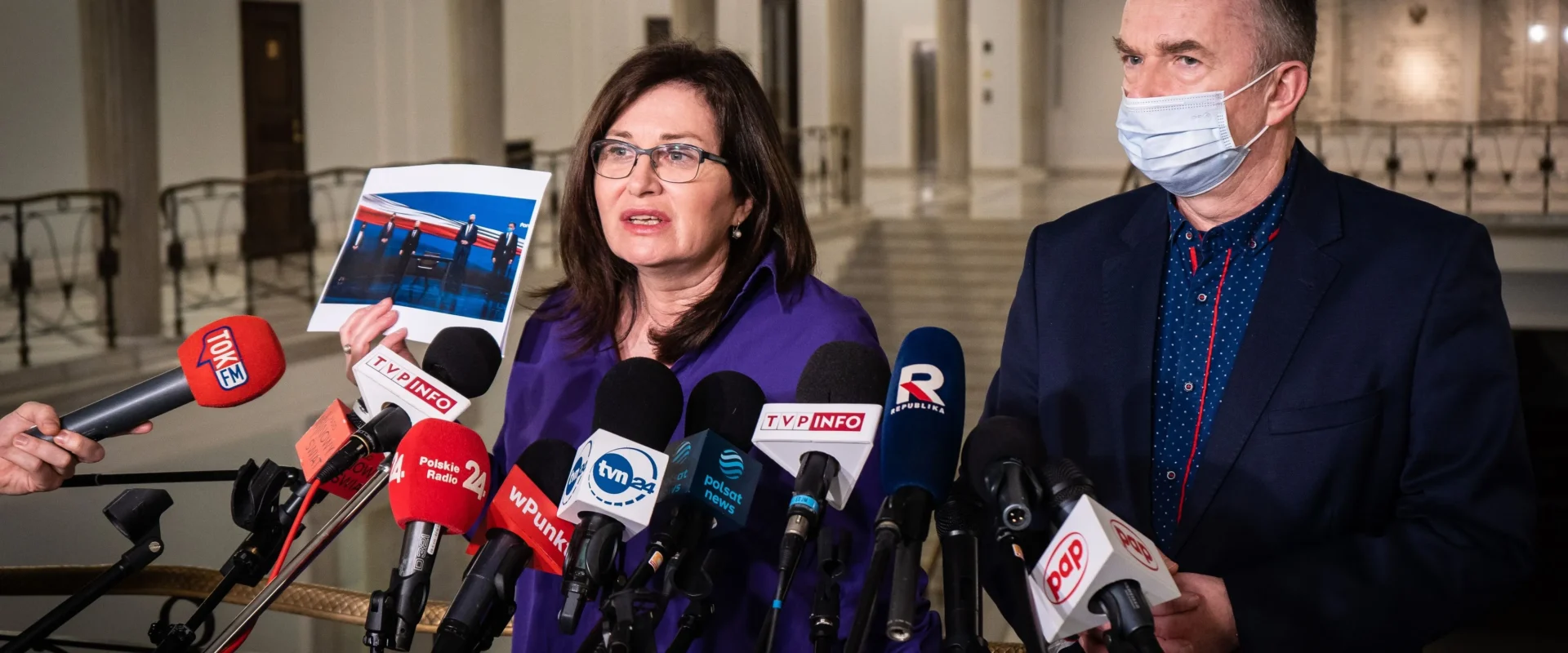 Na zdjęciu widać kobietę prezentującą zdjęcie podczas konferencji prasowej, obok której stoi mężczyzna, a w tle zbiera się grupa dziennikarzy z mikrofonami.