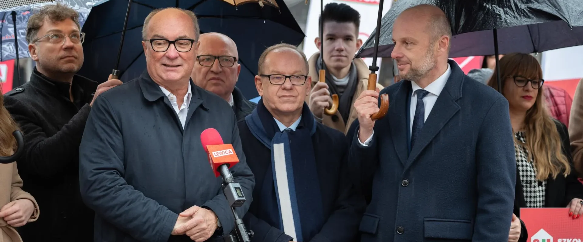 Grupa polityków podczas konferencji prasowej w deszczu, stoją pod parasolami, a przed mikrofonem z logo Lewica przemawia jeden z uczestników.