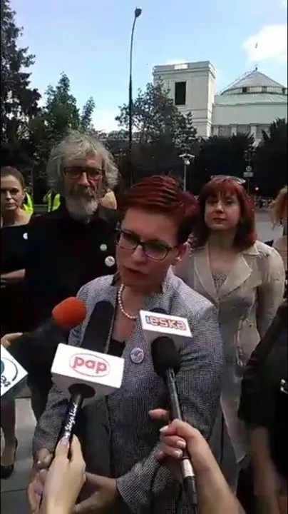 Przedstawicielka Komitetu „Ratujmy Kobiety” udziela wywiadu otoczona mikrofonami i towarzyszącymi osobami na tle budynku.